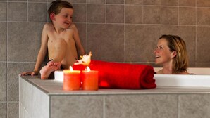Eine Badewanne mit Kind und Erwachsenem, einem roten Handtuch und drei angezündeten Kerzen.