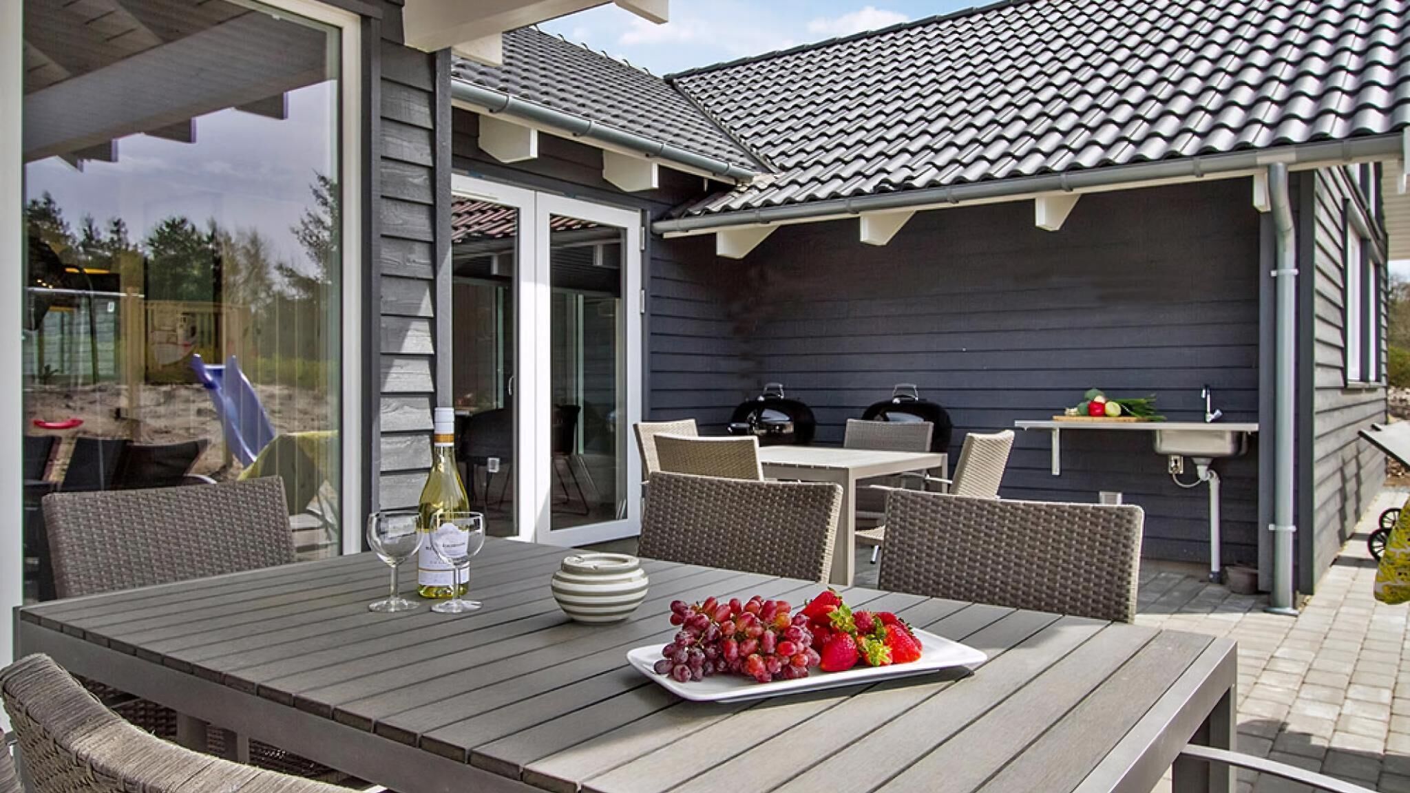 An outdoor patio with a table set for two, a bottle of wine, and a plate of grapes and strawberries.