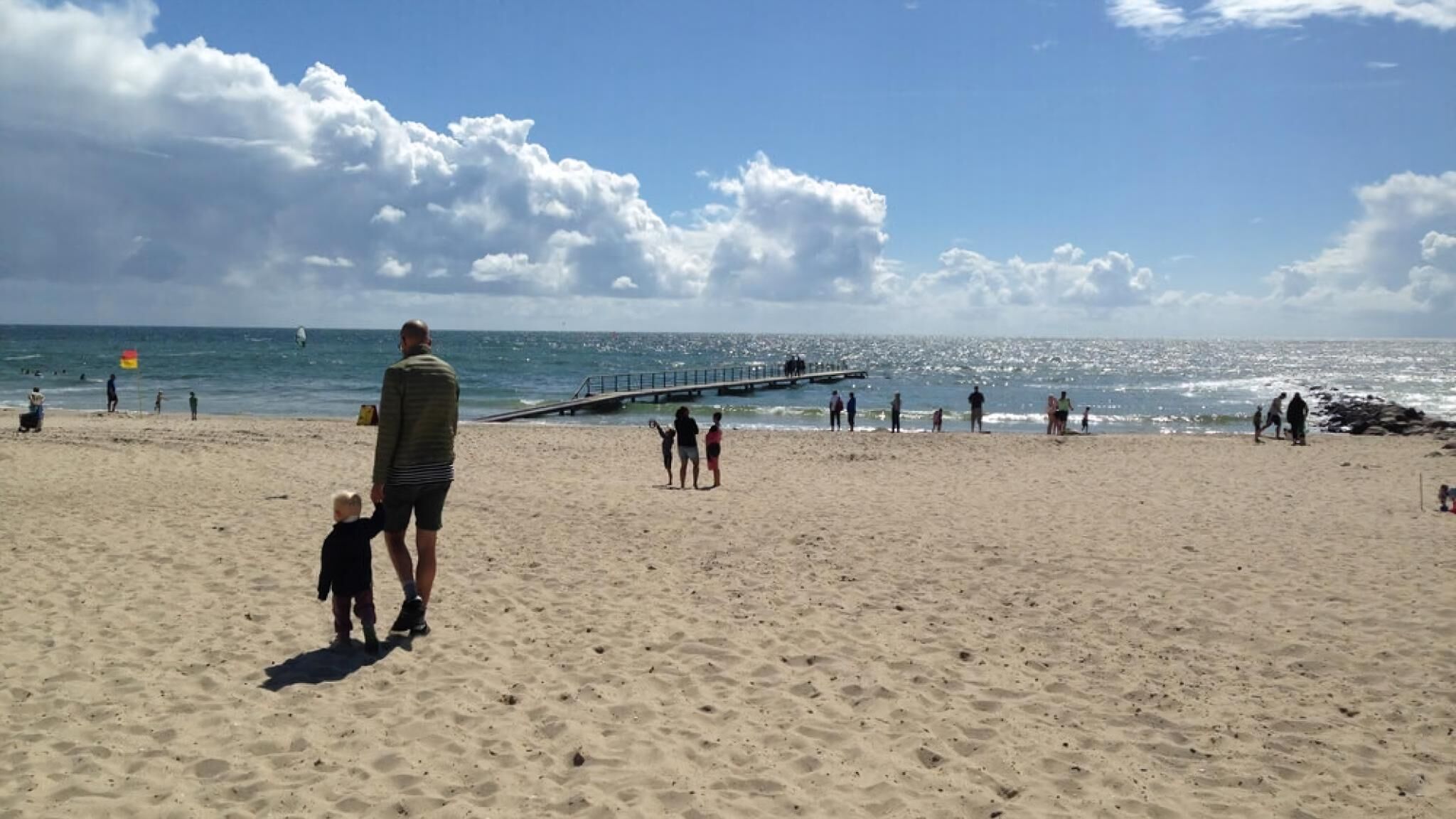 En sandstrand med en mole, der strækker sig ud i vandet, mennesker der går, og en klar himmel.