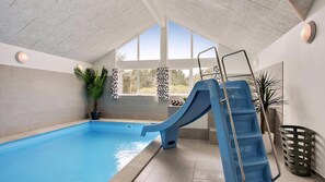An indoor swimming pool with a blue slide, potted plants, and a view of the outdoors through large windows. - VillaVilla 399 - Blåvand, Vestjylland (Blåvand)