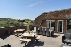 A patio with wooden tables and chairs, a brick wall, and a thatched-roof building with large windows. - VillaVilla 675 - Søndervig, Vestjylland (Ringkøbing)