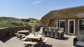 Eine Terrassengaststätte mit Holztischen und -stühlen, ein Gebäude mit einem Reetdach und Blick auf einen Hügel.