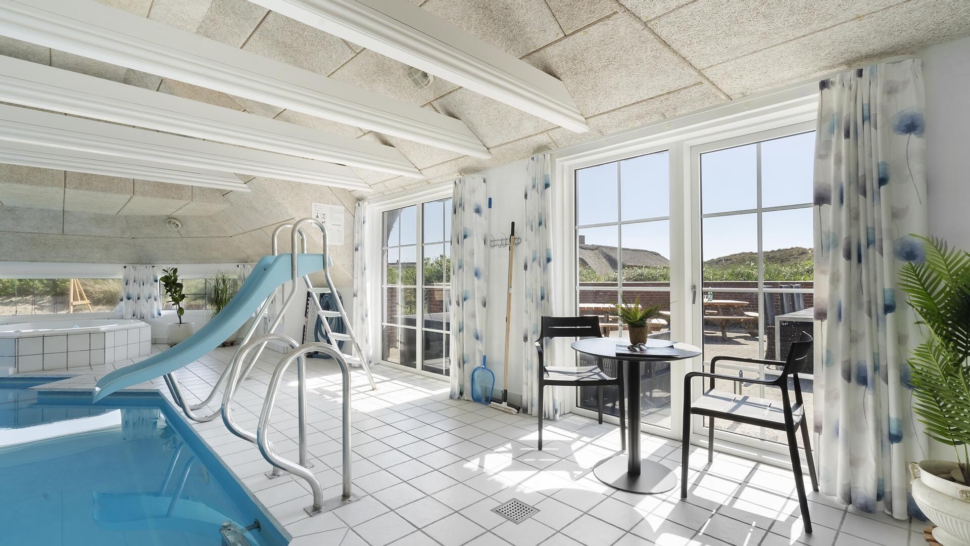 Un área de piscina interior con un tobogán, una terraza con sol con mesa y sillas, y ventanas grandes que ofrecen vistas al exterior.