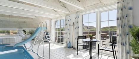 An indoor swimming pool area with a slide, a sun terrace with a table and chairs, and large windows offering a view of the outdoors.