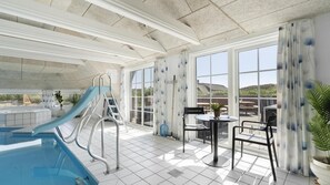 Ein Poolbereich mit Rutsche, eine Sonnenterrasse mit Tisch und Stühlen sowie große Fenster mit Blick ins Freie.