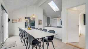 Un espace repas moderne avec une longue table, des chaises noires, des luminaires suspendus et une vue sur la cuisine grâce à une porte vitrée.