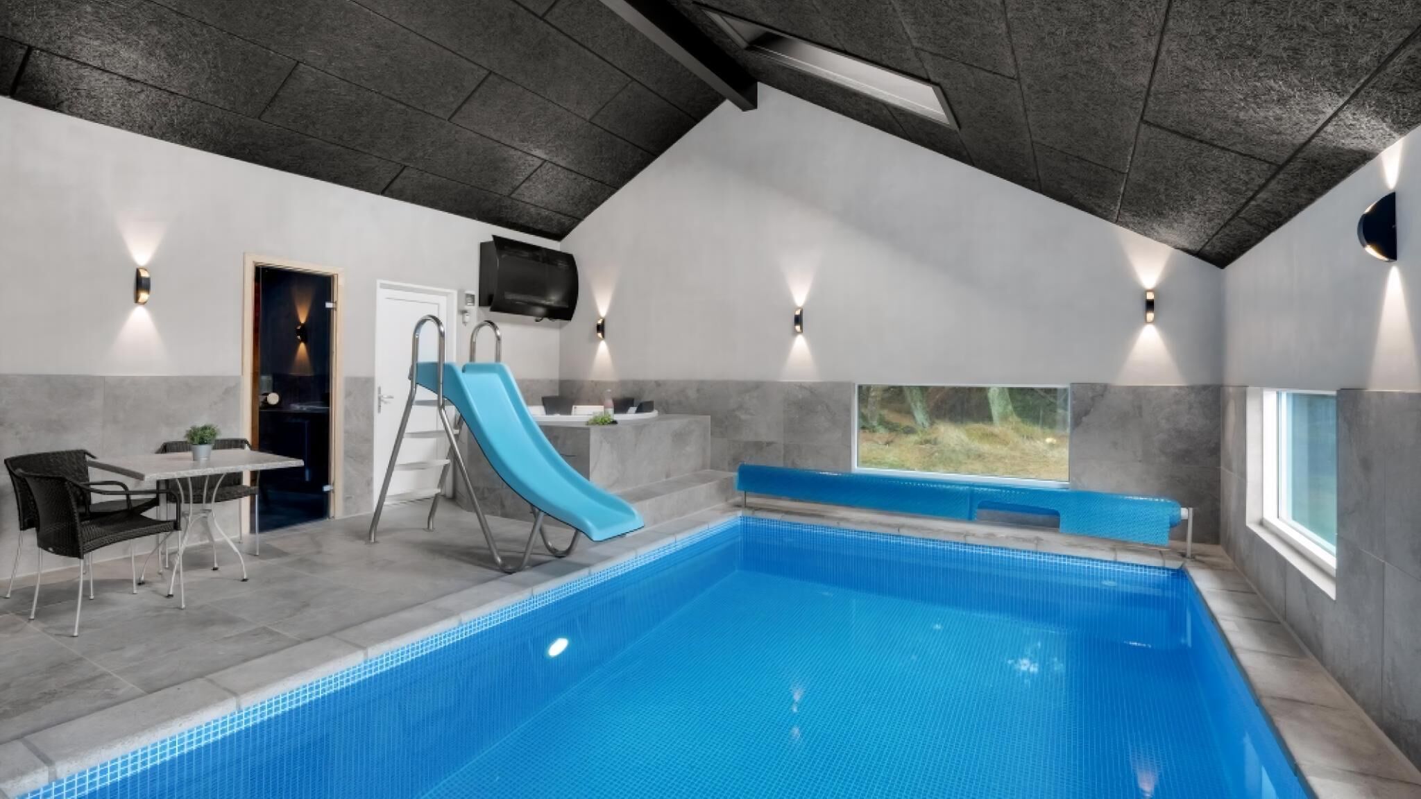 An indoor swimming pool with a blue slide, a table with chairs, and a TV mounted on the wall.