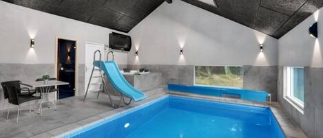 An indoor swimming pool with a blue slide, a table with chairs, and a TV mounted on the wall.