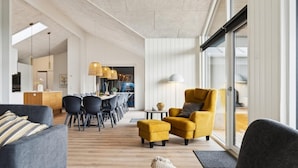 Un soggiorno moderno con un divano, una poltrona e un poggiapiedi, una zona pranzo con sedie e un tavolo, e una cucina sullo sfondo.