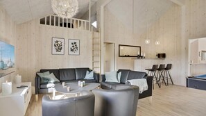 Ein modernes Wohnzimmer mit Sofa, Couchtisch und Sesseln. Es gibt eine Essecke mit einer Bar und Hockern. Der Raum hat Holzwände und einen erhöhten Bereich darüber.
