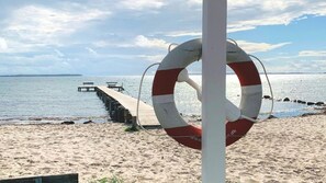 Ein Rettungsring ist an einem Pfahl an einem sandigen Strand befestigt, von wo aus ein Steg ins Wasser führt.