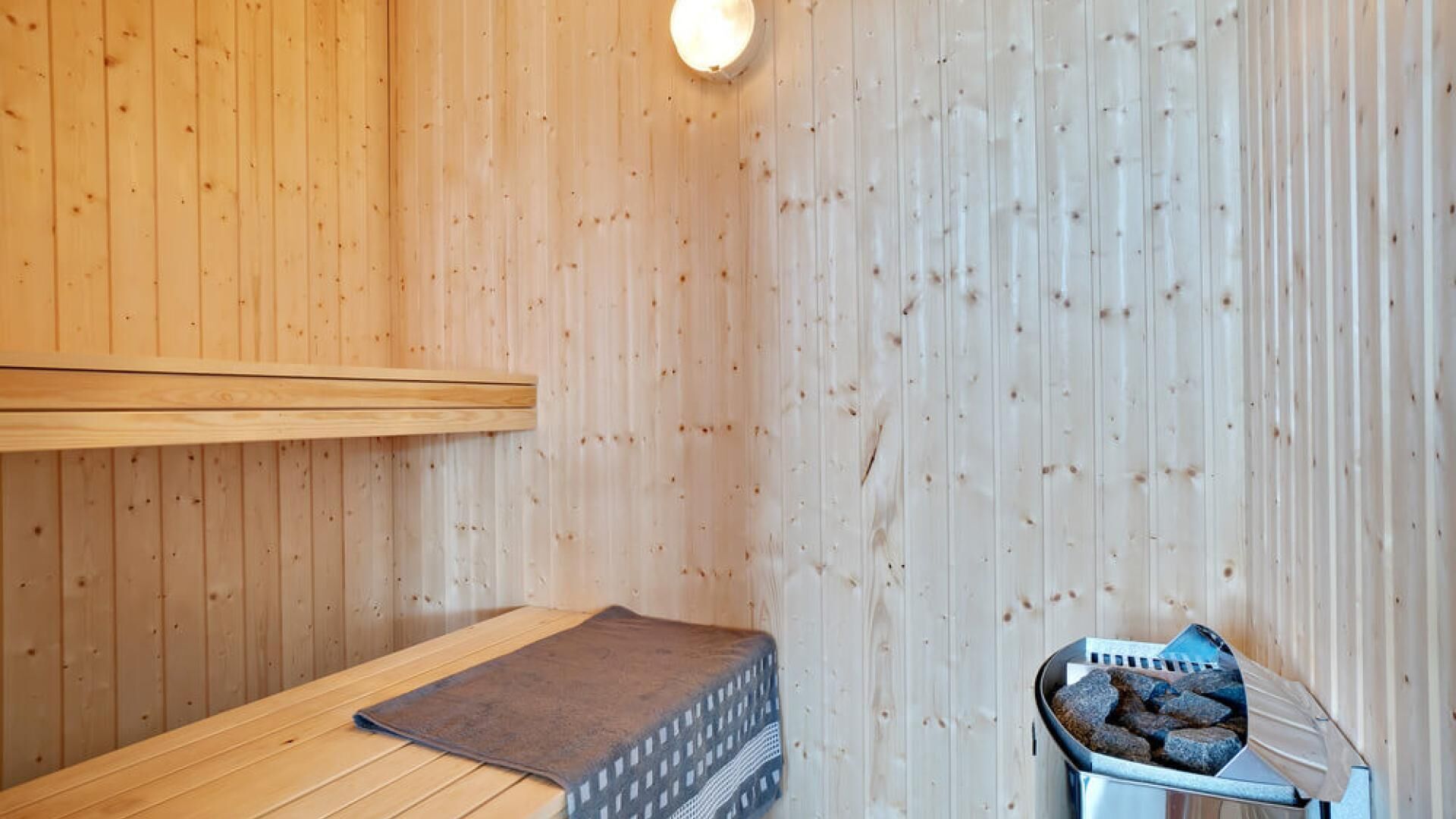 A sauna room with wooden walls, a bench, a mat, and a heater with rocks.