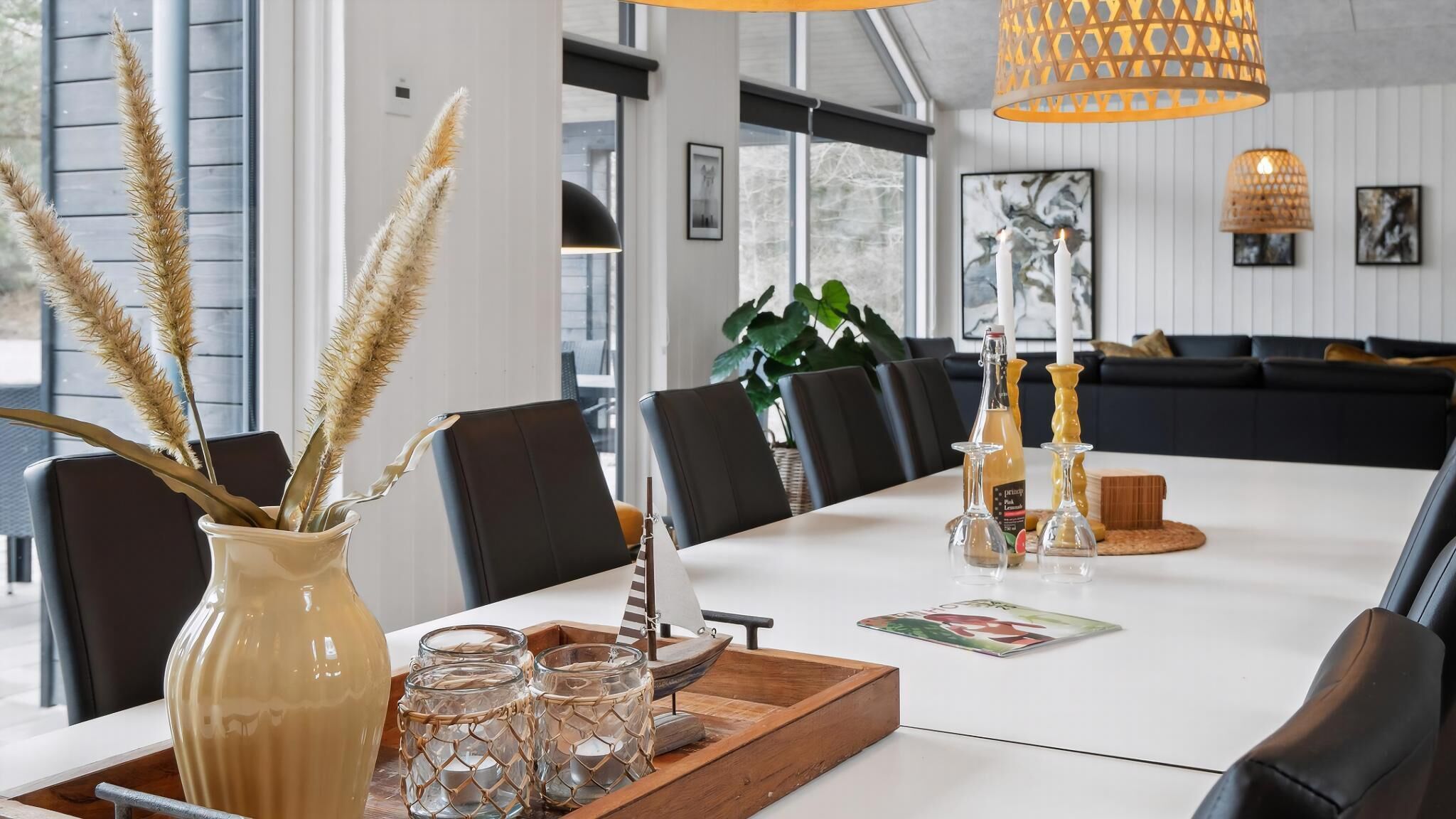 A dining area with a long table, chairs, a vase with dried plants, candle holders, and a bottle.