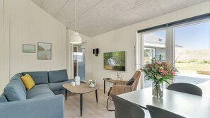 A modern living room with a blue sofa, a coffee table, a TV, and a dining area with chairs and a vase of flowers.