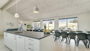 Et rummeligt køkken med en stor ø, moderne pendellamper og udsigt til det fri.