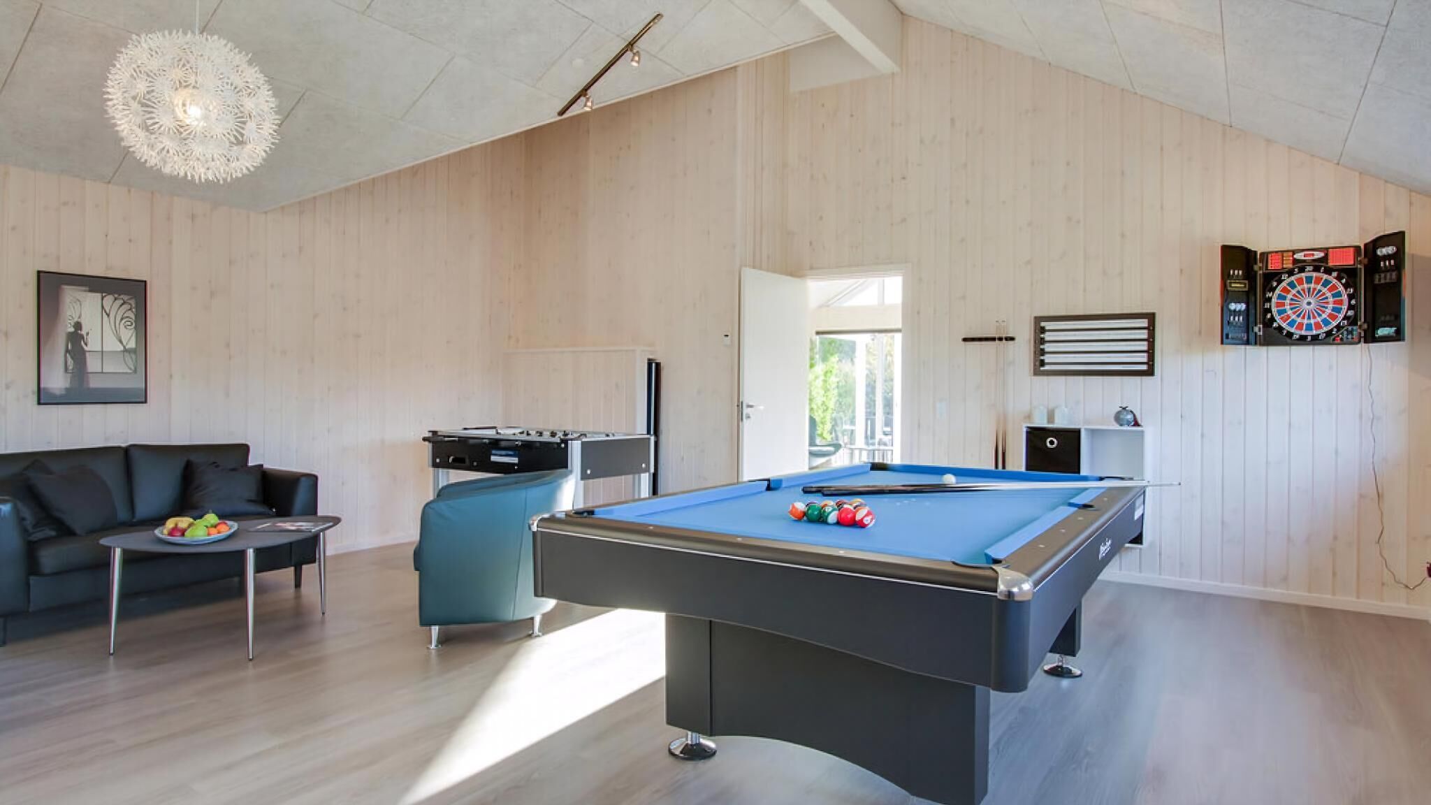 Ein modernes Wohnzimmer mit Billardtisch, einer Couch, einem Couchtisch mit Obst, einer Jukebox und einem Dartboard.