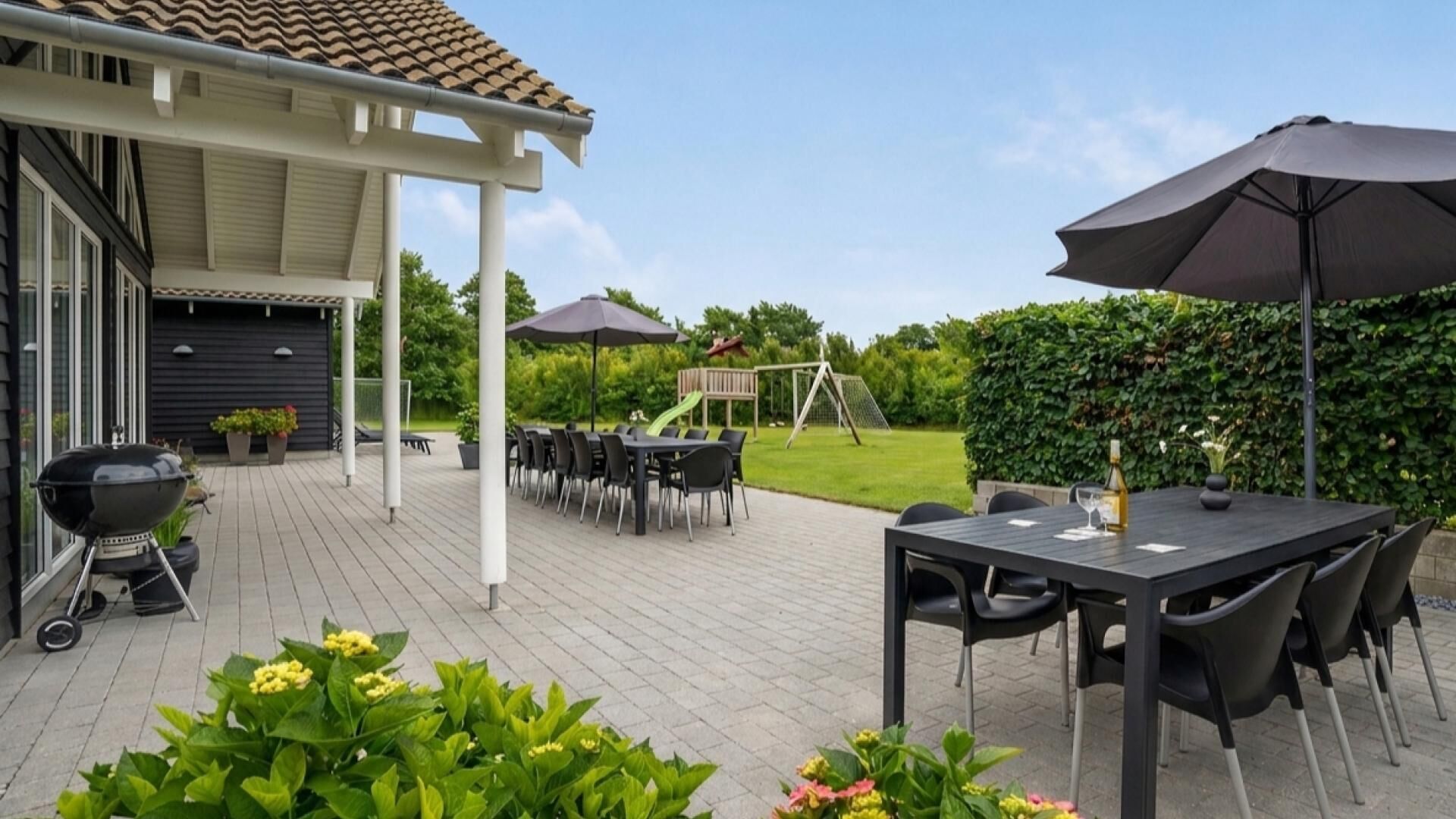 Eine Terrasse im Freien mit Grill, Esstisch und Stühlen, umgeben von Grünpflanzen und einem Spielbereich.