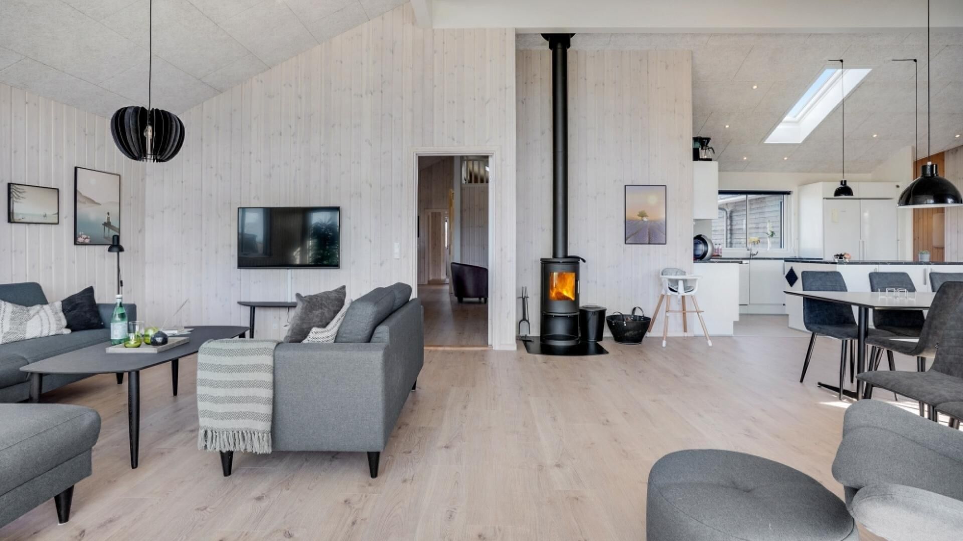 En moderne stue med pejs, grå sofaer, et fjernsyn og et køkkenområde med et spisebord og stole.