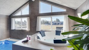 Ein modernes Badezimmer mit einer großen Badewanne, einer Duschkabine und Blick auf ein Haus und die Landschaft durch große Fenster.