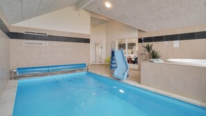 Una piscina interior amb tobogan, zona de dutxes i espai d'estar amb una banca.