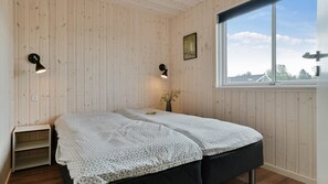 Una habitación con paredes de madera, una cama con colcha blanca estampada, dos lámparas fijadas a la pared, una ventana con vistas y una planta pequeña sobre una mesita de noche.