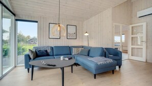 A modern living room with a blue sectional sofa, a round coffee table, and a large window offering a view of the outdoors. - VillaVilla 578 - Marielyst, Lolland/Falster/møn (Idestrup)