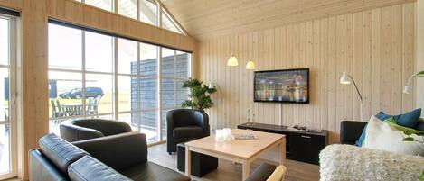 A modern living room with a wooden ceiling, large windows, a black leather sofa, a wooden coffee table, and a TV mounted on the wall.