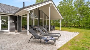 Een overdekt terras met buitenmeubilair, een betegelde vloer en uitzicht op een huis met grote ramen.