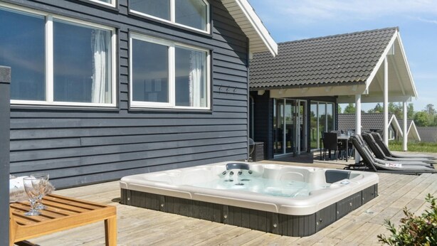 Una casa moderna con un amplio jacuzzi en una terraza de madera, ventanas grandes y una zona de estar cubierta al aire libre.