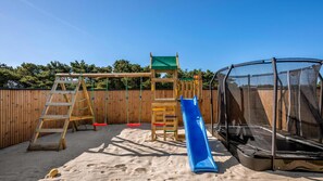 A wooden play structure with a slide, a ladder, and a climbing area, surrounded by a sandy area and a wooden fence. - VillaVilla 332 - Søndervig, Vestjylland (Ringkøbing)