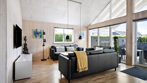Een moderne woonkamer met een zwarte bank, houten vloeren, een aan de muur bevestigde televisie en grote ramen.
