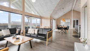 A spacious living area with a black leather sofa, wooden coffee table, and a dining table with chairs. Large windows provide natural light and views of the outdoors. - VillaVilla 322 - Skåstrup Strand, Fyn og øer (Bogense)