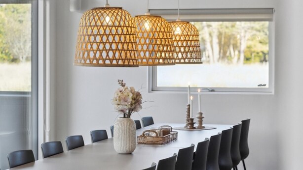 Een eethoek met een witte tafel, zwarte stoelen, een geweven hanglamp, een vaas met bloemen en kaarsen.
