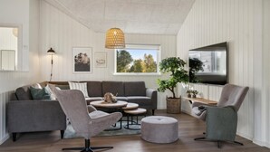 Een moderne woonkamer met een grijze bank, twee fauteuils, een salontafel en een aan de muur bevestigde televisie.