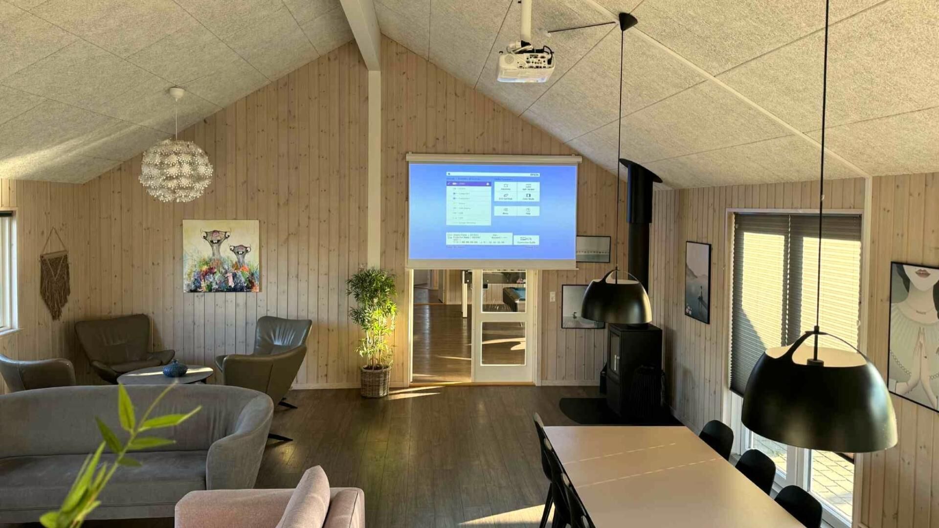 A spacious living area with a large screen displaying a presentation, wooden walls, a modern chandelier, and a cozy seating arrangement.