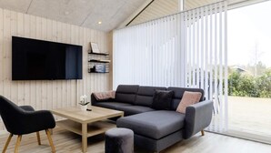 A modern living room with a sectional sofa, a wooden coffee table, and a black armchair. A flat-screen TV is mounted on the wall, and there are shelves with books and decorative items. - VillaVilla 650 - Marielyst, Lolland/Falster/møn (Væggerløse)