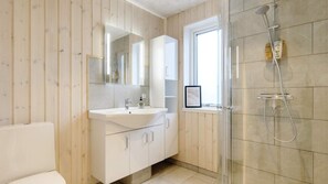 Un baño moderno con un panel de pared de madera, un lavabo blanco con un espejo arriba, una cabina de ducha de vidrio y una ventana que proporciona luz natural.