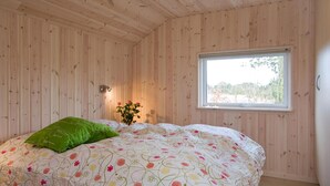 Ein Schlafzimmer mit Holzwänden, einem Bett mit floralem Bettbezug, einem grünen Kopfkissen, einer Nachttischlampe und einem Fenster mit Blick auf Bäume.