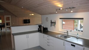 Eine moderne Küche mit weißen Schränken, einer Edelstahl-Spüle und einem Geschirrspüler. Ein Fenster bietet Blick ins Freie, an der Wand hängt eine Uhr und über den Schränken ist ein Fernseher angebracht.