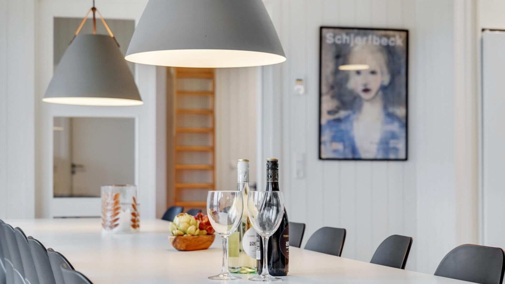 A dining area with a white table, two wine bottles, and glasses. There are two pendant lights above the table. A poster with the name 'Schjortbeck' is on the wall.