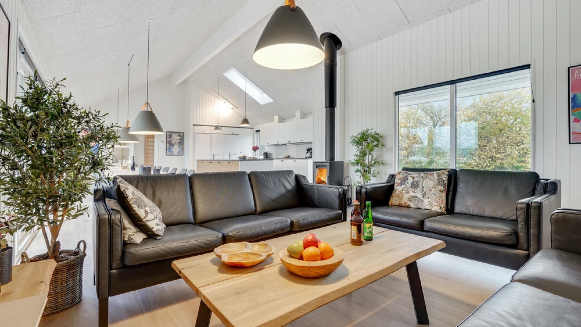 A modern living room with leather sofas, a wooden coffee table with fruit and bottles, and a large window with a view of trees.