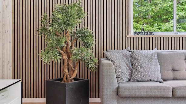 Een moderne woonkamer met een grijze bank, gestreepte kussens en een grote plant in pot voor een wand van houten latten.