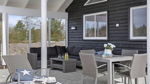 A covered outdoor seating area with wicker furniture, a table with a vase of flowers, and a dark wooden building in the background. - VillaVilla 393 - Blåvand, Vestjylland (Blåvand)