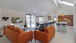 A modern living room with orange sofas, a dining area, a kitchen with a bar, and a large TV. - VillaVilla 393 - Blåvand, Vestjylland (Blåvand)