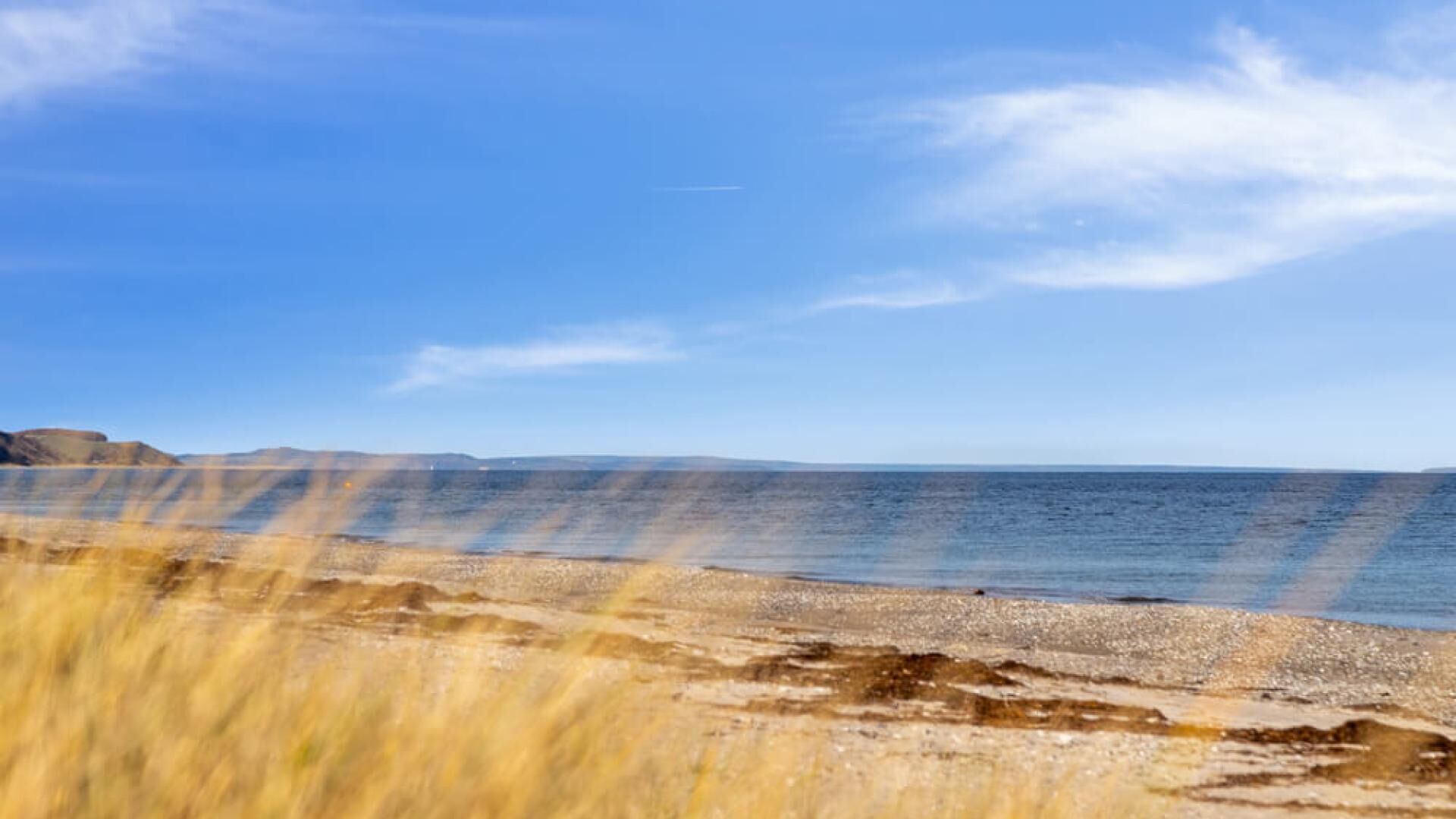 Un paysage côtier avec un ciel dégagé, une plage de sable et des collines à l’horizon.