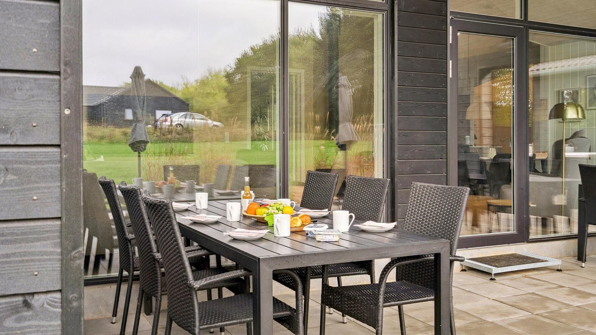 An outdoor dining area with a table set for four, surrounded by wicker chairs, and large windows offering a view of a green landscape.