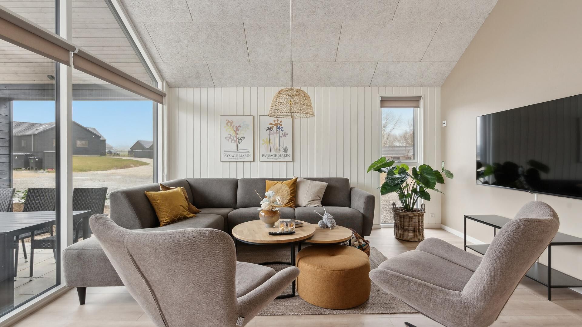 A modern living room with a grey sofa, two armchairs, a coffee table, and a TV. There are two framed artworks on the wall, a large window, and a view of the outside.