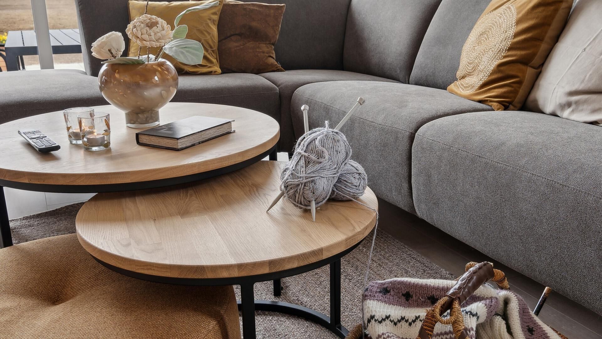 A modern living room with a grey sofa, a round wooden coffee table, a vase with flowers, and a knitted blanket.