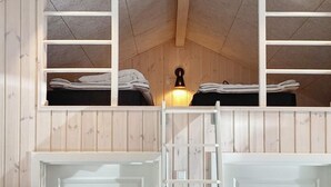 Um quarto pequeno e acolhedor com paredes de madeira e teto inclinado. Há duas camas com cobertores e travesseiros bem arrumados, um banco branco e um aplique na parede.
