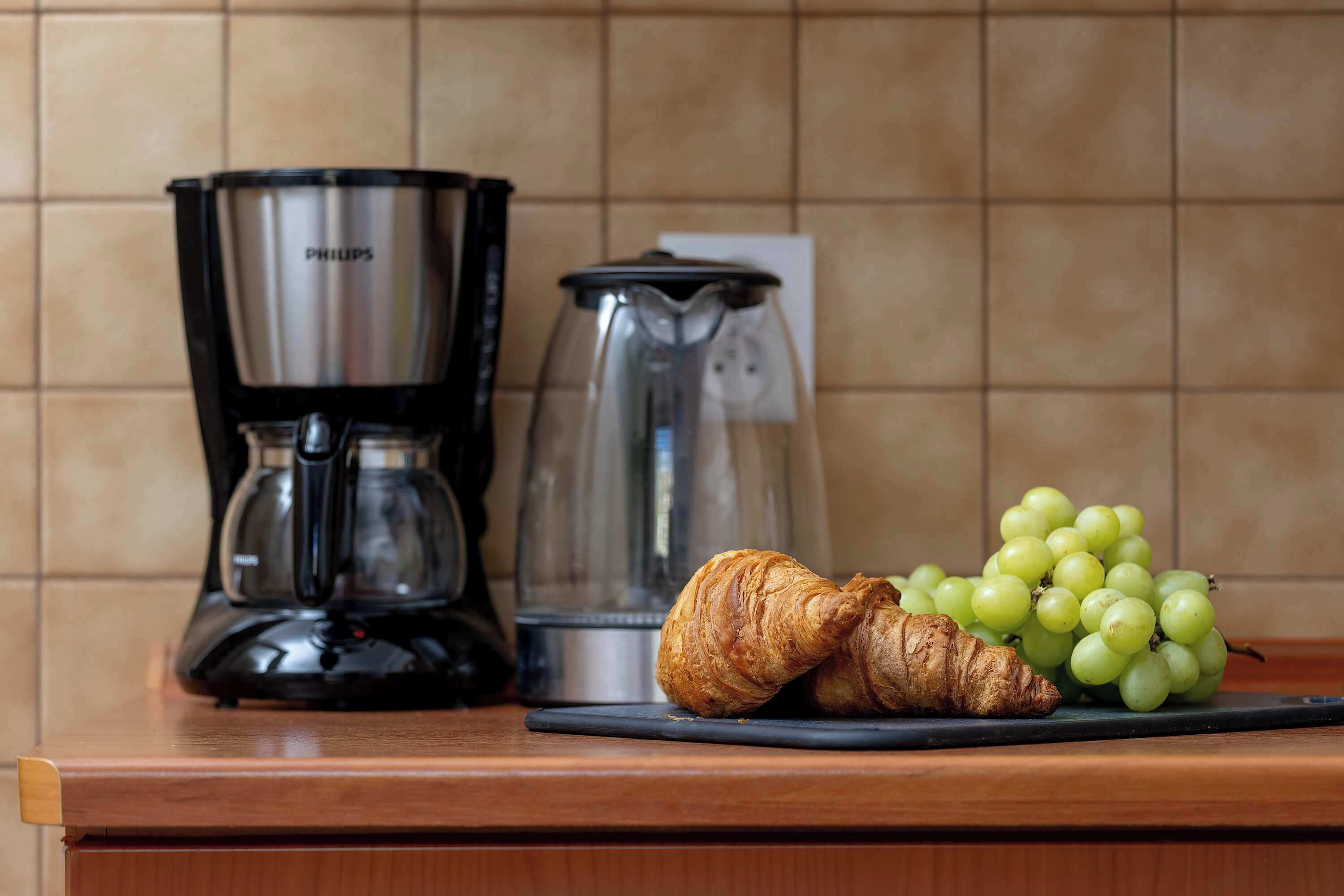 Una encimera de cocina con una cafetera, una jarra de vidrio, un croissant y un racimo de uvas.